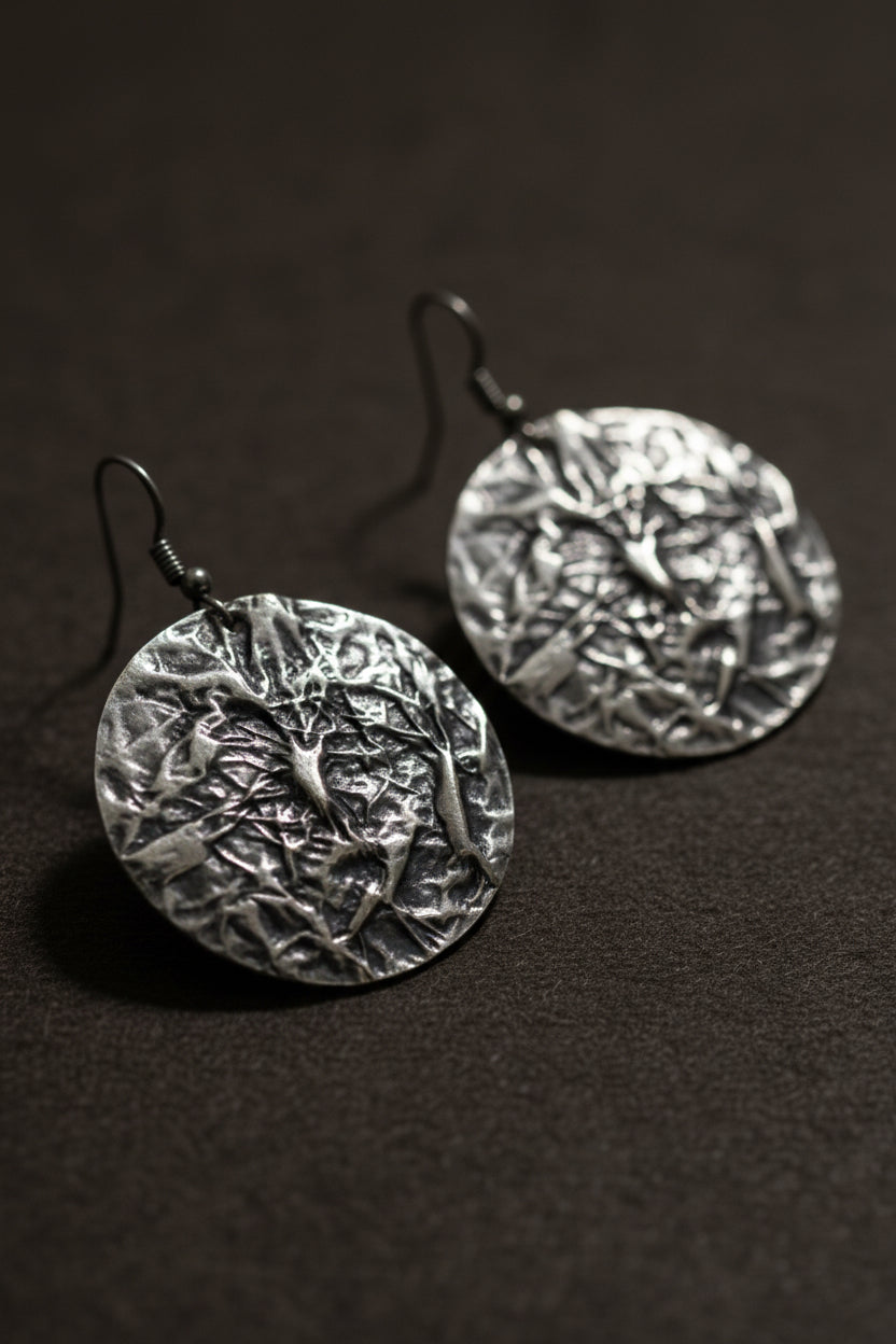 A premium 3D photorealistic render hero shot of a single Aura Textured Silver Disc Earring, suspended against a sophisticated soft gradient grey background. The close-up view emphasizes the deep, complex crumpled metal foil texture of the large silver circular disc, with dramatic studio lighting creating brilliant highlights and shadows across the multi-faceted surface.