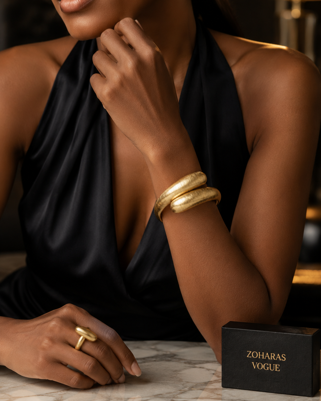 A close-up lifestyle shot of  model wearing Aurora_Statement_Bracelet__ZoharasVogue_and a matching gold ring, paired with a black satin halter top. A black jewelry box with the brand name 'ZOHARAS VOGUE' is visible on a marble surface in the foreground.he