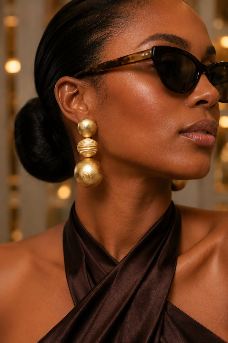 Model wearing Calia Matte Sculpted Drops Earrings_ZoharasVogue, large gold beaded drop earrings, and a dark brown draped halter top. The background features warm, out-of-focus golden bokeh lights.