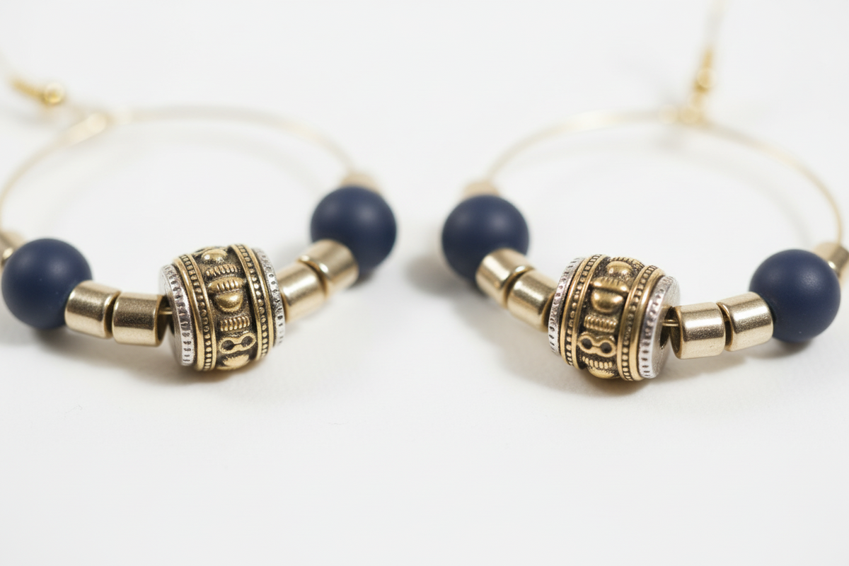 Gold wire hoop earrings with matte navy beads, ornate gold barrel bead, and gold spacer beads, shown on a textured neutral surface.
