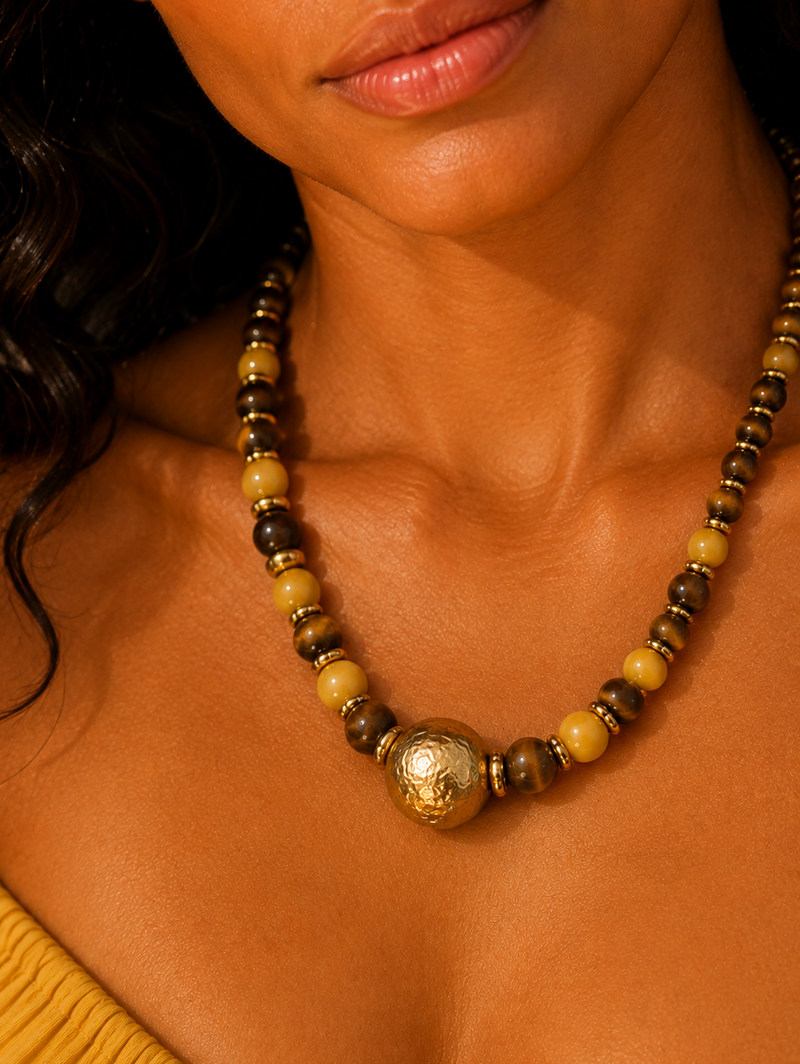 A close-up of a model wearing a beaded necklace with round yellow, brown, and metallic gold beads, featuring a prominent textured gold pendant at the center. By ZoharasVogue 
