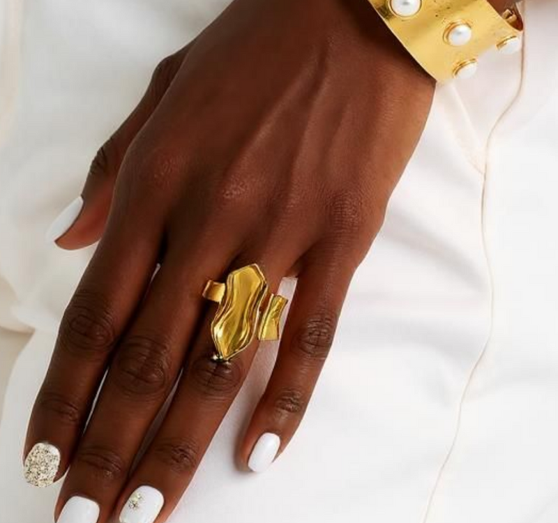 Close-up of a woman’s hand wearing the Aurora Flow 18K Gold Statement Ring with an abstract molten gold design, paired with a gold and pearl cuff bracelet. ZoharasVogue 