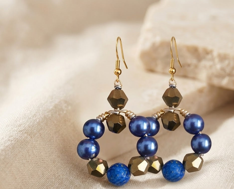 A close-up high-impact portrait of a woman wearing a single intricate gold wire-wrapped hoop earring, identical in design to the earrings The earring features spherical cobalt blue pearl beads and natural Lapis Lazuli stone mixed with geometric faceted metallic gold beads. The woman's skin has a natural glow in the warm light. Text overlay reads "Statement Gold: The Art of the Bold Earring. ZoharasVogue