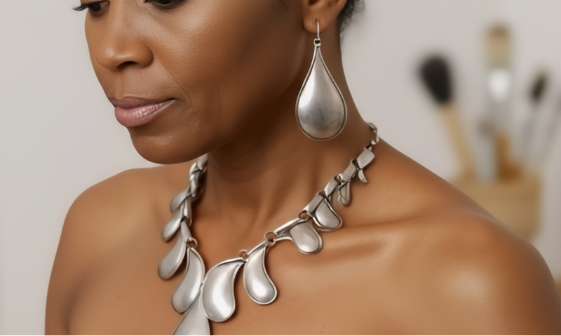 Woman in a black dress with a silver necklace and earrings, standing in front of a colorful abstract painting. ZoharasVogue