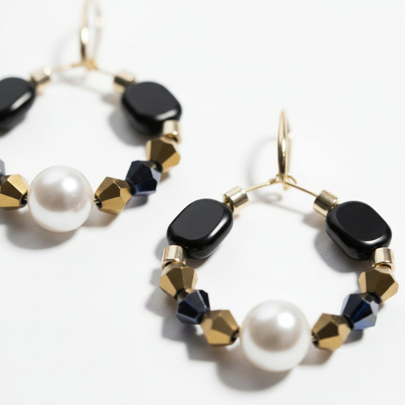 Gold hoop earrings featuring a central pearl, black geometric beads, navy crystal accents, and gold faceted spacer beads on a white background. ZoharasVogue
