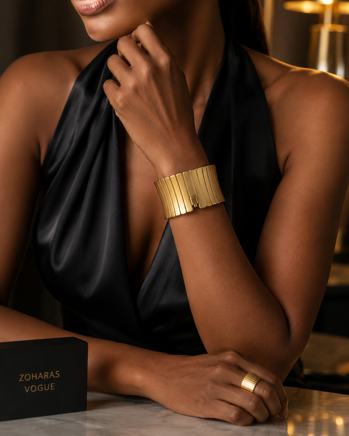 A woman in a black satin halter top wearing Monarque Gleam Bracelet - ZOHARAS VOGUE v4 and a matching gold ring, with a black Zoharas Vogue box on a marble surface in the foreground.