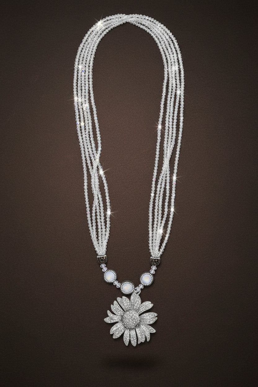 A long multi-strand beaded necklace with a silver-toned flower-shaped pendant and small dark spacer beads, laid flat on a beige textured surface. ZoharasVogue