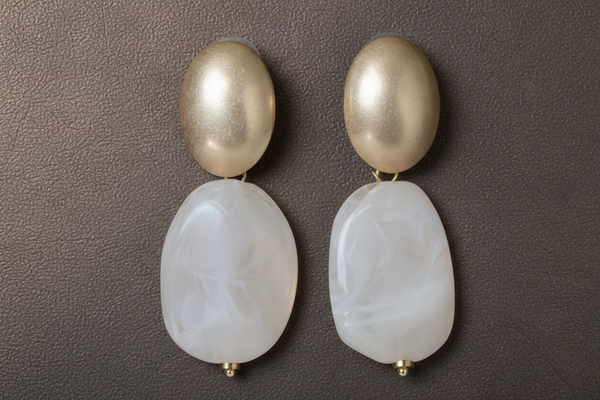 A pair of drop earrings with a matte gold oval top and a large, teardrop-shaped marbled cream stone hanging below, displayed on a beige textured background. ZoharasVogue