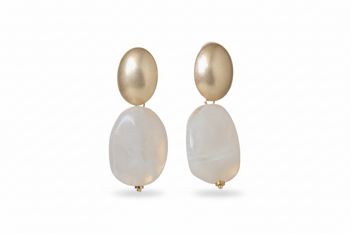 A pair of drop earrings with a matte gold oval top and a large, teardrop-shaped marbled cream stone hanging below, displayed on a beige textured background. ZoharasVogue
