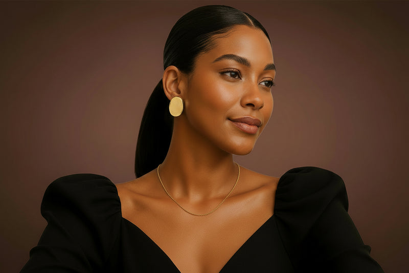 Model is wearing gold sculptured earrings with a black top with puffed sleeves against a beige background