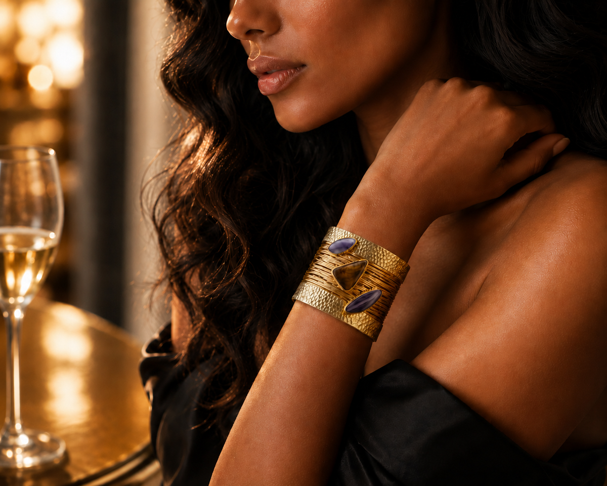 The The_Obsidian_Empress_Cuff_ZoharasVoguezv, a close-up lifestyle shot of a woman wearing a wide gold cuff bracelet with textured bands and two dark purple stone inlays. She is wearing a black off-the-shoulder top, and a glass of white wine is visible on a gold table in the softly lit background.
