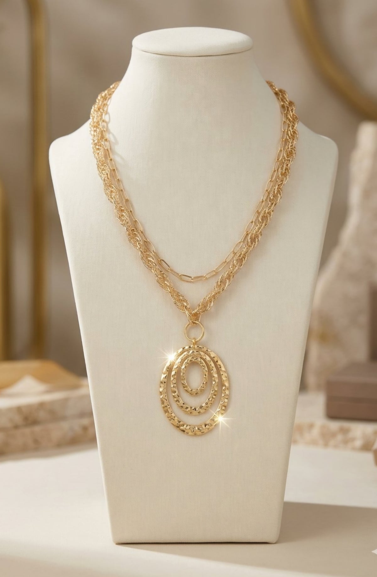 A gold layered necklace with a textured spiral pendant, displayed on a white jewelry bust against a softly blurred boutique background. ZoharasVogue 