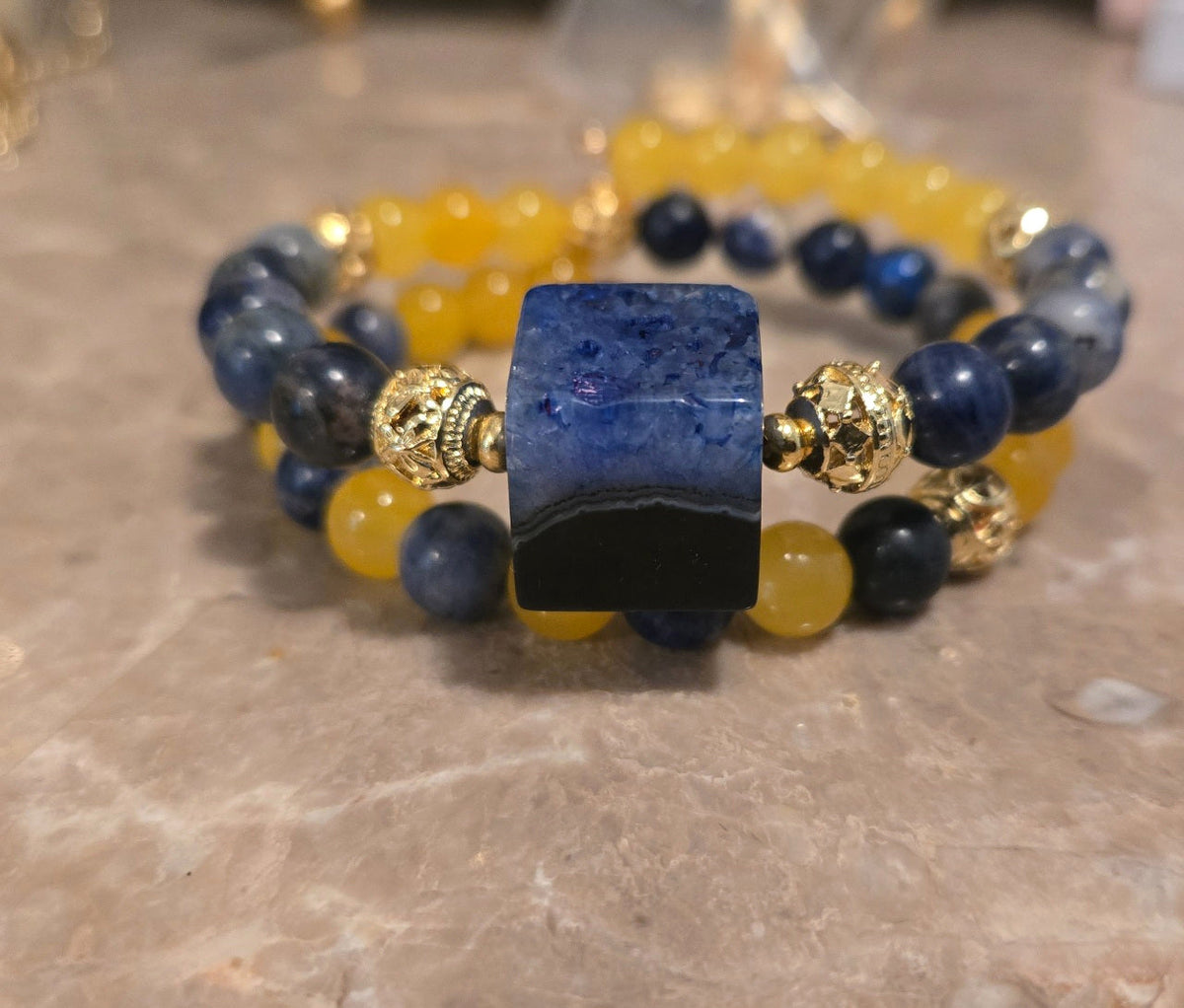 Real product photo of Azure Solstice Bracelet: two-row blue and yellow beaded stretch bracelet with gold details and marbled cube.
