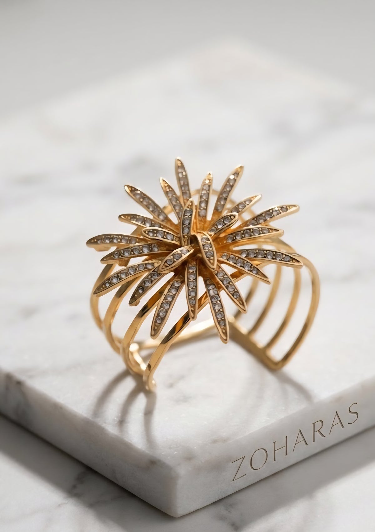 Gold bracelet with star-like design on a marble surface with 'Zoharas' branding. A close-up of a person's wrist wearing a wide, multi-layered gold brass cuff bracelet with a hammered texture, showcasing four parallel bands.