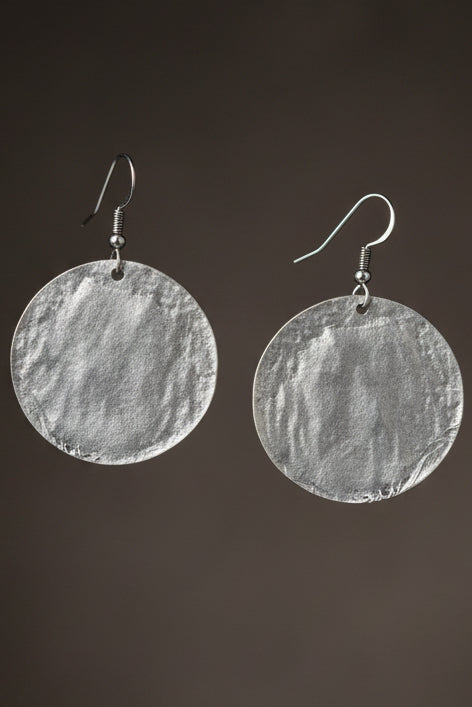 A premium 3D photorealistic render hero shot of a single Aura Textured Silver Disc Earring, suspended against a sophisticated soft gradient grey background. The close-up view emphasizes the deep, complex crumpled metal foil texture of the large silver circular disc, with dramatic studio lighting creating brilliant highlights and shadows across the multi-faceted surface.